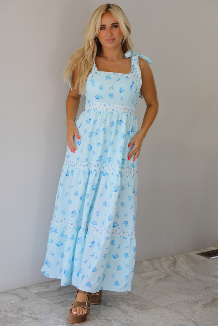 Woman wearing a light blue floral maxi dress indoors against a white wall with white marble flooring. 