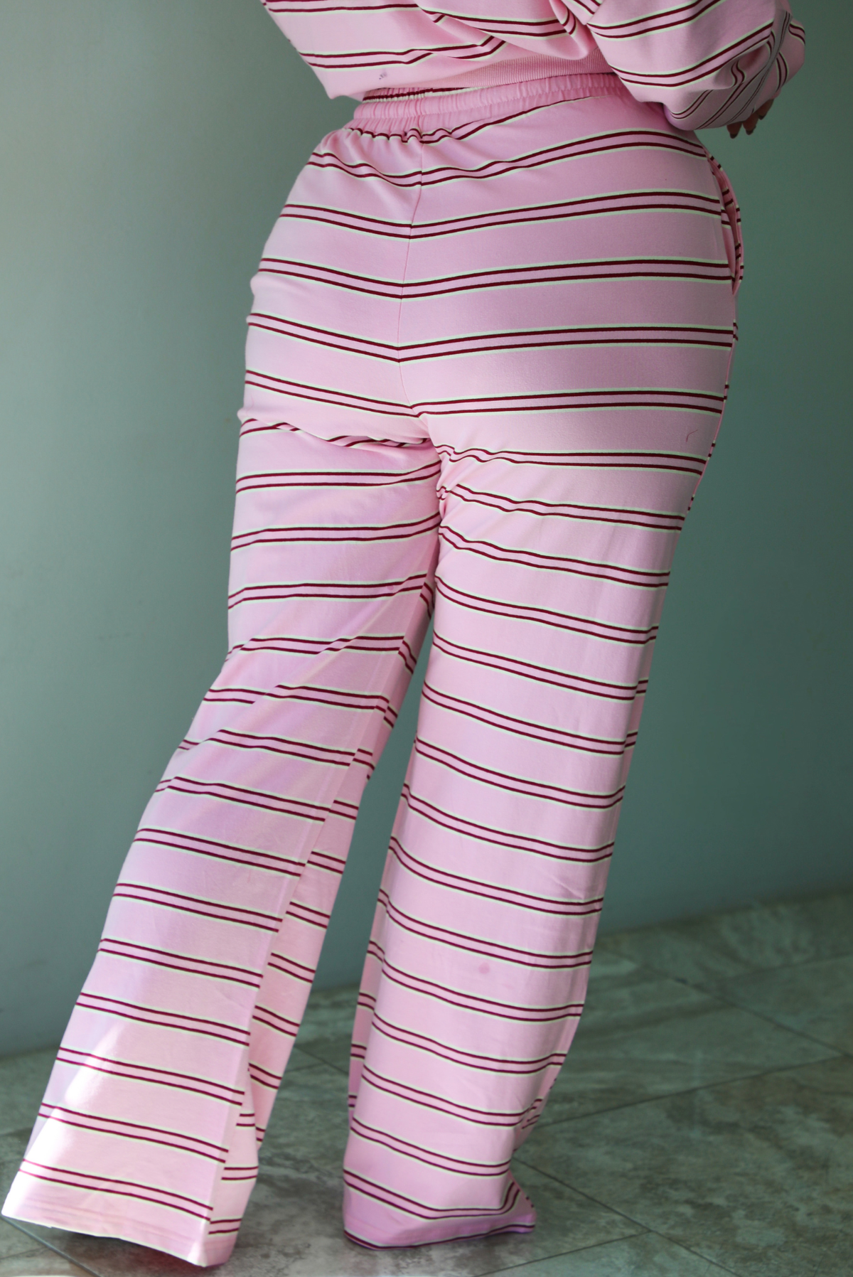 A woman shown from behind wearing a pink two piece sweat set featuring a maroon and white striped design on a sweatshirt and high waisted, wide leg pants, standing indoors against a white wall with white marble flooring. The photo shows a closeup of the pants, which have an elastic and drawstring waistline and functional pockets on either side. 