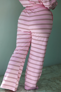 A woman shown from behind wearing a pink two piece sweat set featuring a maroon and white striped design on a sweatshirt and high waisted, wide leg pants, standing indoors against a white wall with white marble flooring. The photo shows a closeup of the pants, which have an elastic and drawstring waistline and functional pockets on either side. 