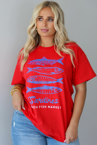 A woman wearing a red graphic t-shirt with blue sardine illustrations and the text 'Sardines LISBON FISH MARKET', paired with blue jeans, standing indoors against a white wall.