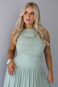 Woman wearing a sage green maxi length dress, which has a rouched detail at the bust, a fitted waistline, a flowy skirt. The photo is taken indoors against a white wall and shows a closeup of the bodice of the dress. 