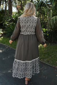 Back of a woman model wearing an olive and off-white stitched detail dress outdoors. Dress has a corset-appearance bodice, long sleeves, functional pockets on either side, an elastic waistline, and a long, maxi length.