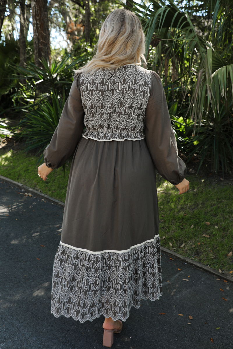 Back of a woman model wearing an olive and off-white stitched detail dress outdoors. Dress has a corset-appearance bodice, long sleeves, functional pockets on either side, an elastic waistline, and a long, maxi length.