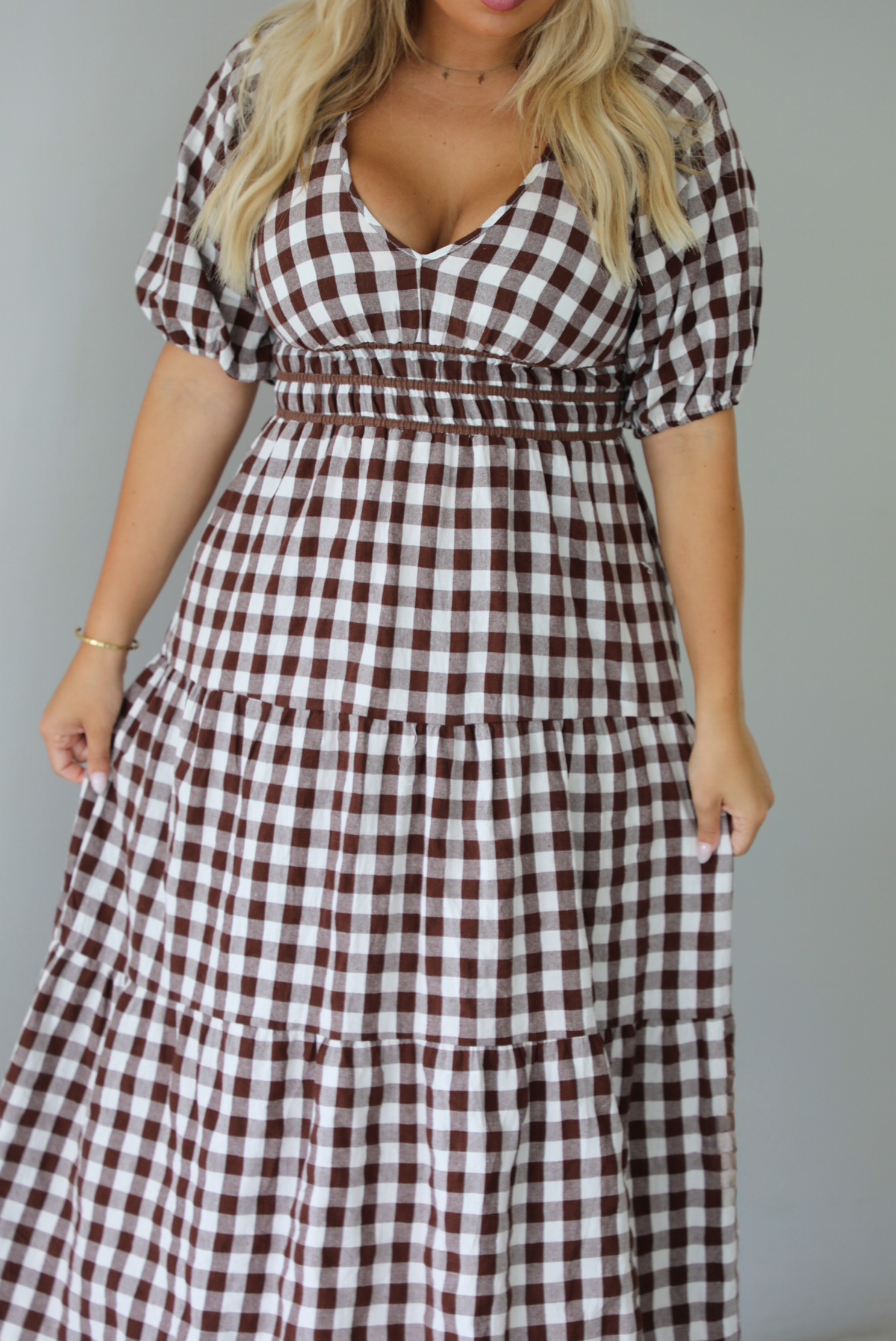 A woman modeling a brown and white gingham tiered midi dress with puff sleeves and a smocked waist, paired with beige slide sandals, standing indoors against a white wall.