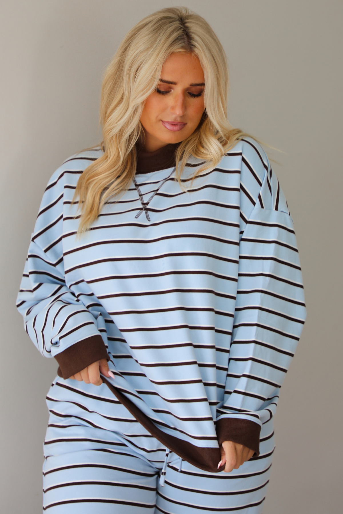 A woman wearing a blue matching two piece set with dark brown and white striped details, featuring an oversized long sleeved top and high waisted loose fitting, flared leg pants, indoors against a white wall. The model has the top untucked, showing the full length, wth the brown hem detail on both the bottom hem of the top and the wrists. 