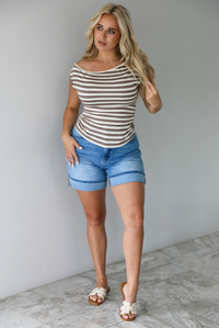 Woman wearing a mocha and white striped top with an off the shoulder design with medium wash denim shorts indoors against a white wall with white marble flooring. 