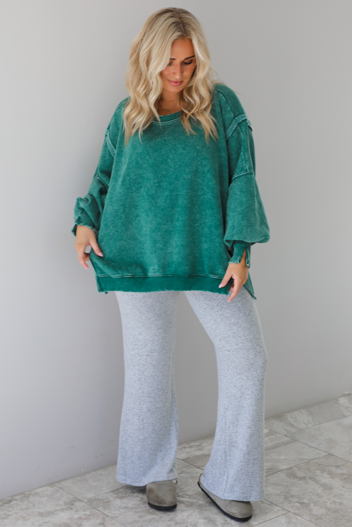Woman wearing green colored oversized sweatshirt with exposed seam details and a high-low design with heather grey casual pants. The outfit is styled with grey slip on shoes. The photo is taken indoors against a white wall with white marble flooring. 