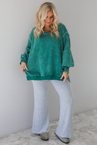 Woman wearing green colored oversized sweatshirt with exposed seam details and a high-low design with heather grey casual pants. The outfit is styled with grey slip on shoes. The photo is taken indoors against a white wall with white marble flooring. 