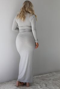 Woman wearing a grey two piece set featuring a turtle neck long sleeved fitted top and high waisted maxi length fitted skirt. The photo shows the back of the set. The photo is taken indoors against a white wall with white marble flooring. 