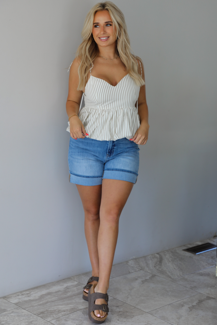 Woman wearing a white and taupe striped tank with medium wash denim shorts indoors against a white wall with white marble flooring. 
