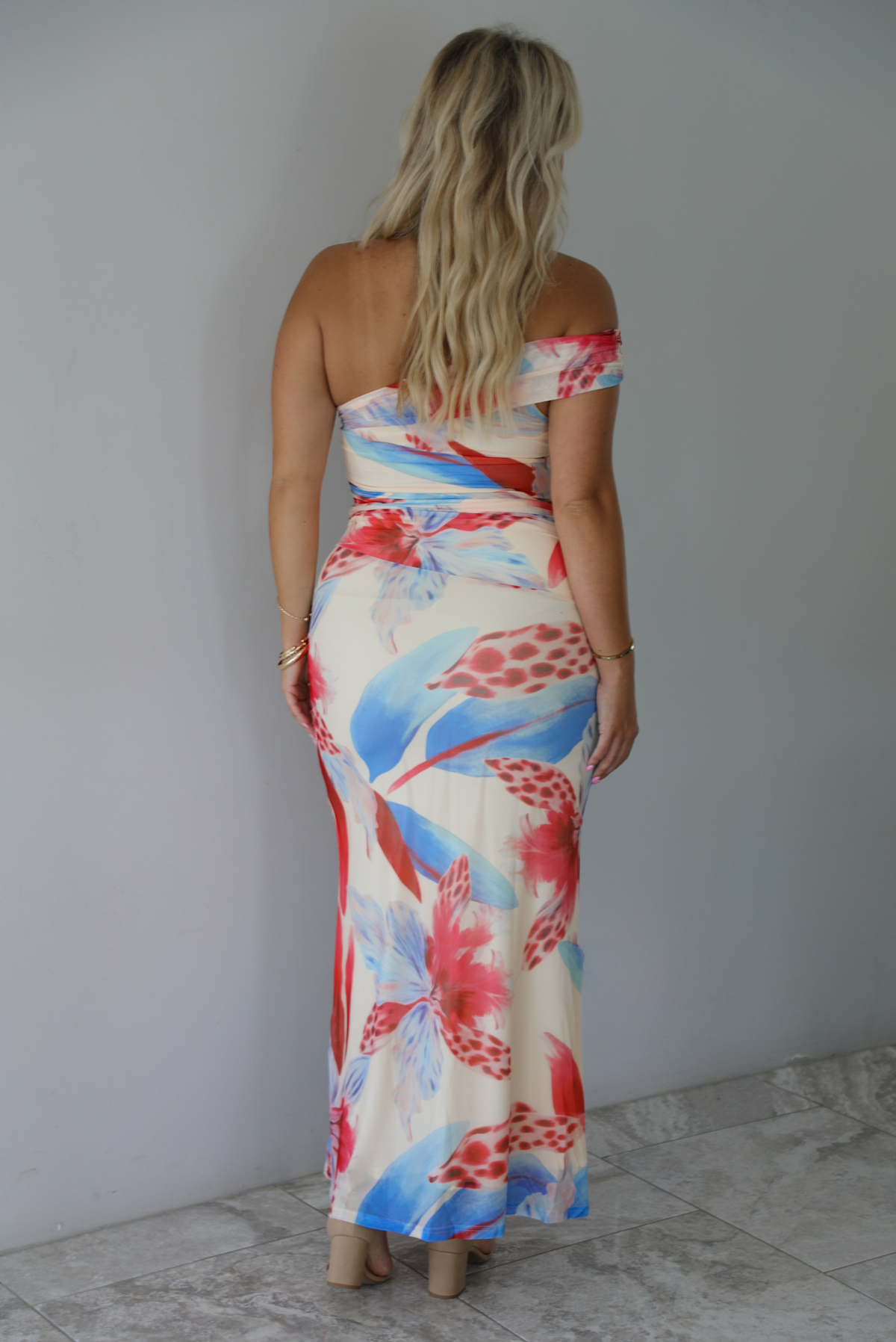 A woman modeling a strapless off-the-shoulder maxi dress with a vibrant red, blue, and cream floral print against a white wall with white marble flooring. The photo shows the back of the dress. 