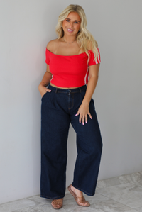 A blonde woman modeling a bright red off-the-shoulder short-sleeve top with white stripes on the sleeves, paired with dark wide-leg denim jeans, standing indoors against a white wall with white marble flooring. 