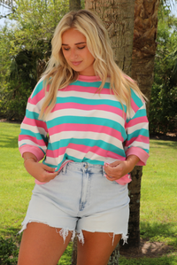 Woman wearing turquoise, pink, and white striped top with light wash denim shorts outdoors with greenery and palm trees in the background. 