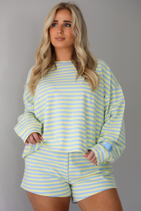 Woman wearing a blue and pale yellow striped two piece set featuring an oversized sweatshirt and shorts. The photo is taken indoors against a white wall and shows a closeup of the set, which is untucked to show the full length.