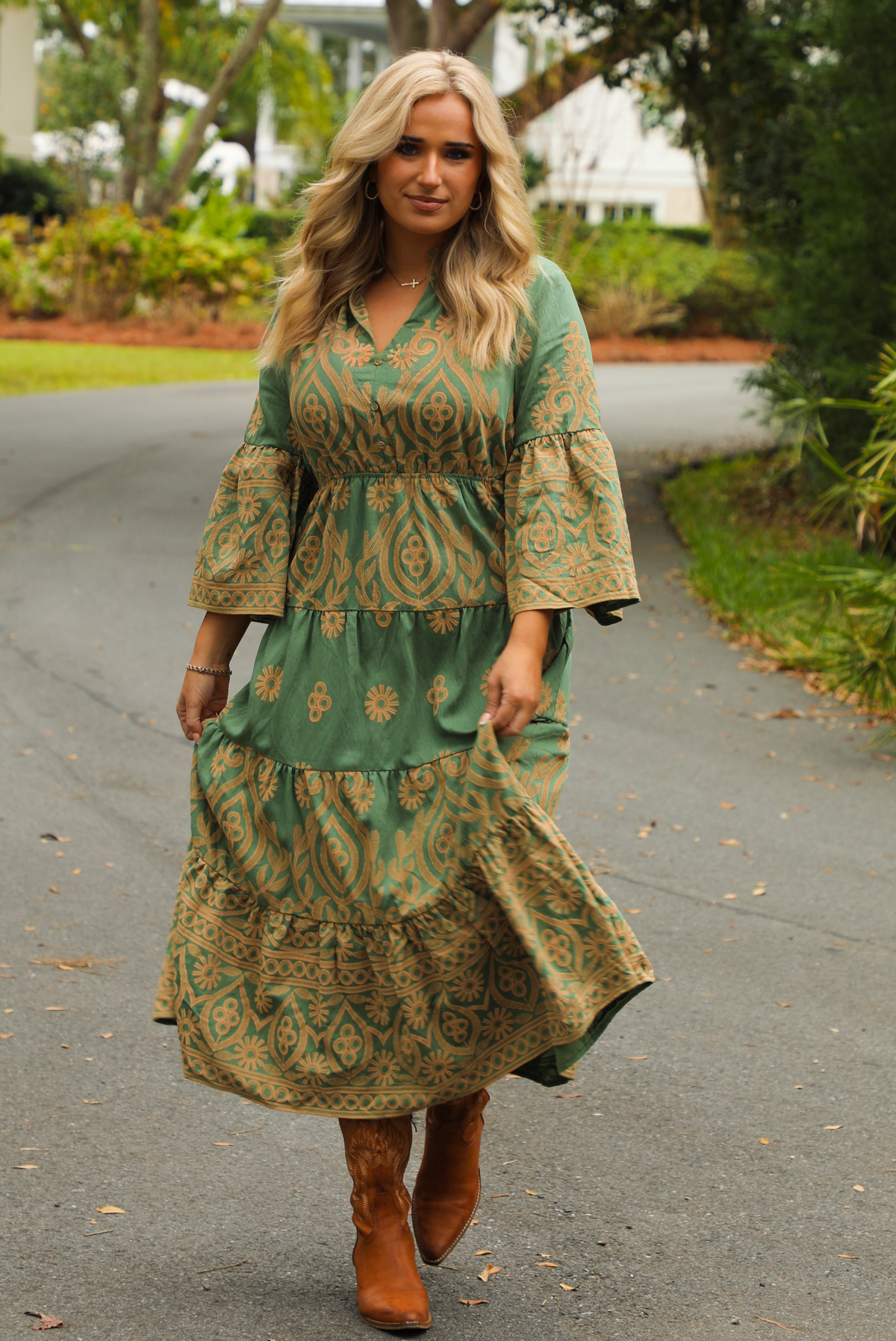 Woman wearing a teal dress that has tan a stitched pattern throughout, quarter length  sleeves, a collared, v-neckline, cinched with elastic waistline, and a maxi-length skirt. The photo is taken outdoors standing on a paved road with trees and other greenery in the background.