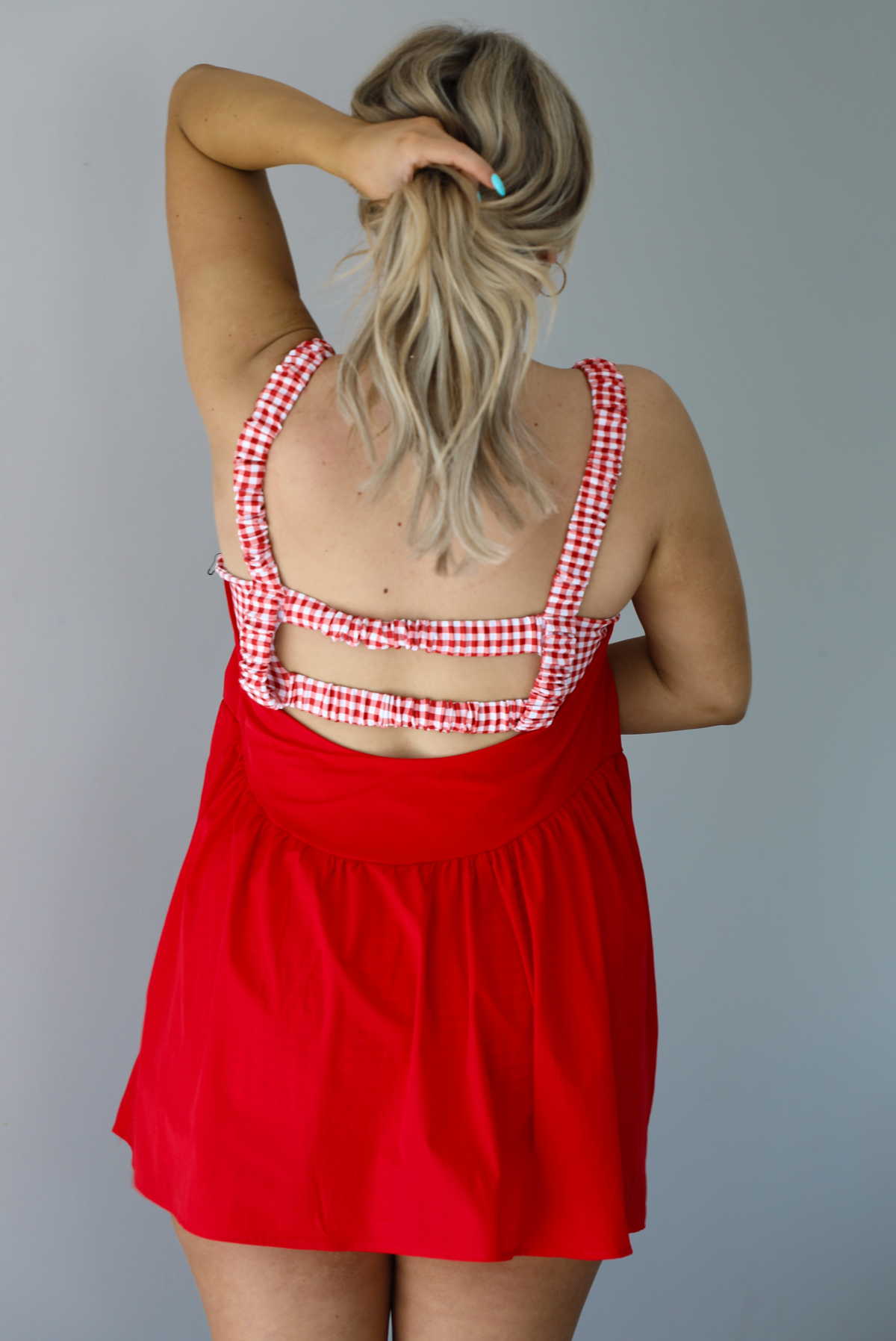 Woman wearing a red dress with a red and white gingham romper underneath. The photo is taken indoors against a white wall and shows the back of the dress, which has elastic double straps going cross the back.