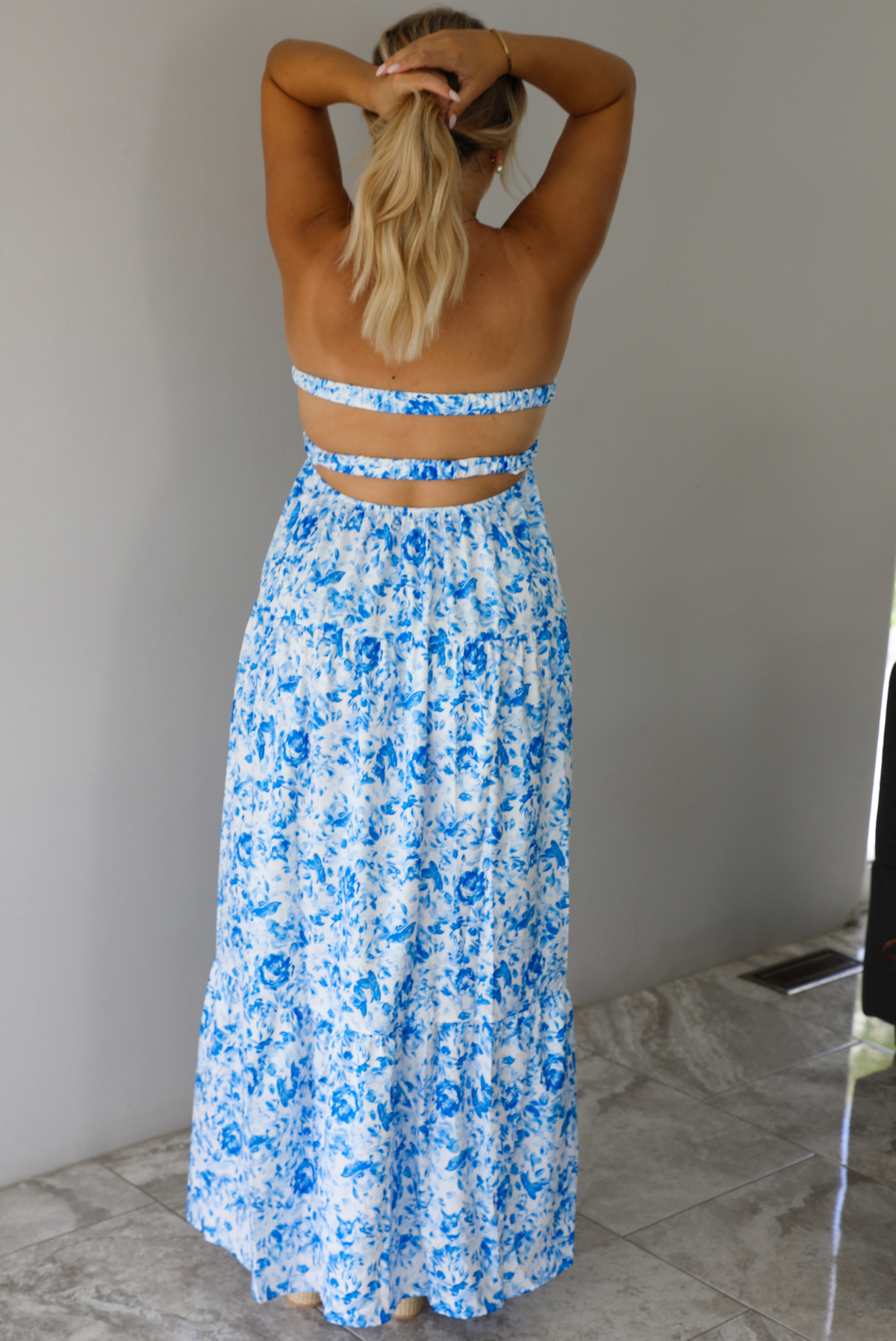 A woman shown from behind modeling a strapless white maxi dress with a blue floral print and a sweetheart neckline with a small keyhole cutout, standing indoors against a white wall with white marble flooring. 