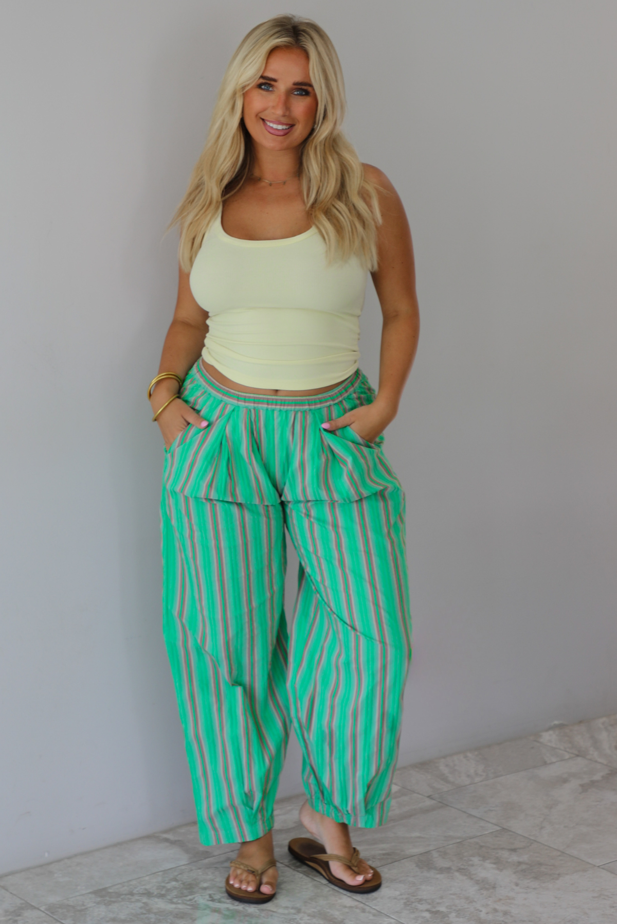 A woman wearing a pale yellow tank and mid-rise pants that have a green, brown, lime green, and tan striped pattern, which have an elastic waistline, functional pockets on either side, and cinched details at the ankles, standing indoors against a white wall with white marble flooring. 