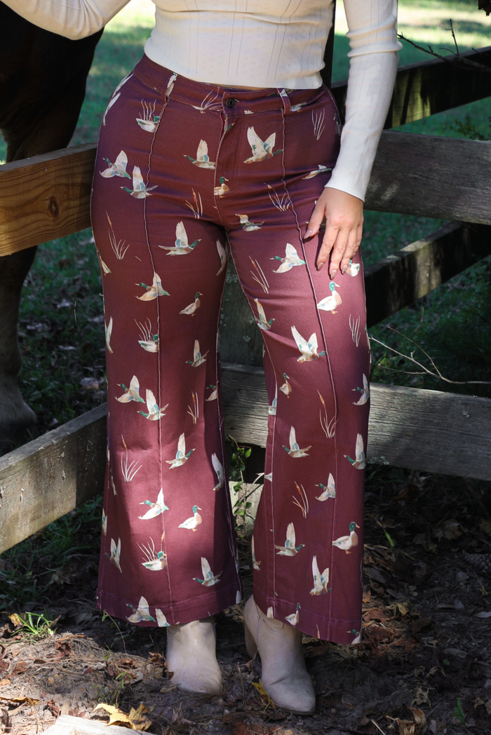 A closeup of a woman wearing a pair of maroon denim pants with a multi-colored repeating mallard design throughout have have a high waisted design, pleated details on the front, standing in front of a wooden fence with greenery and a horse leg in the background. 