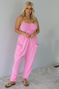 Woman wearing a pink strapless jumpsuit with a fitted bodice and slouchy pants indoors against a white wall with white marble flooring. 