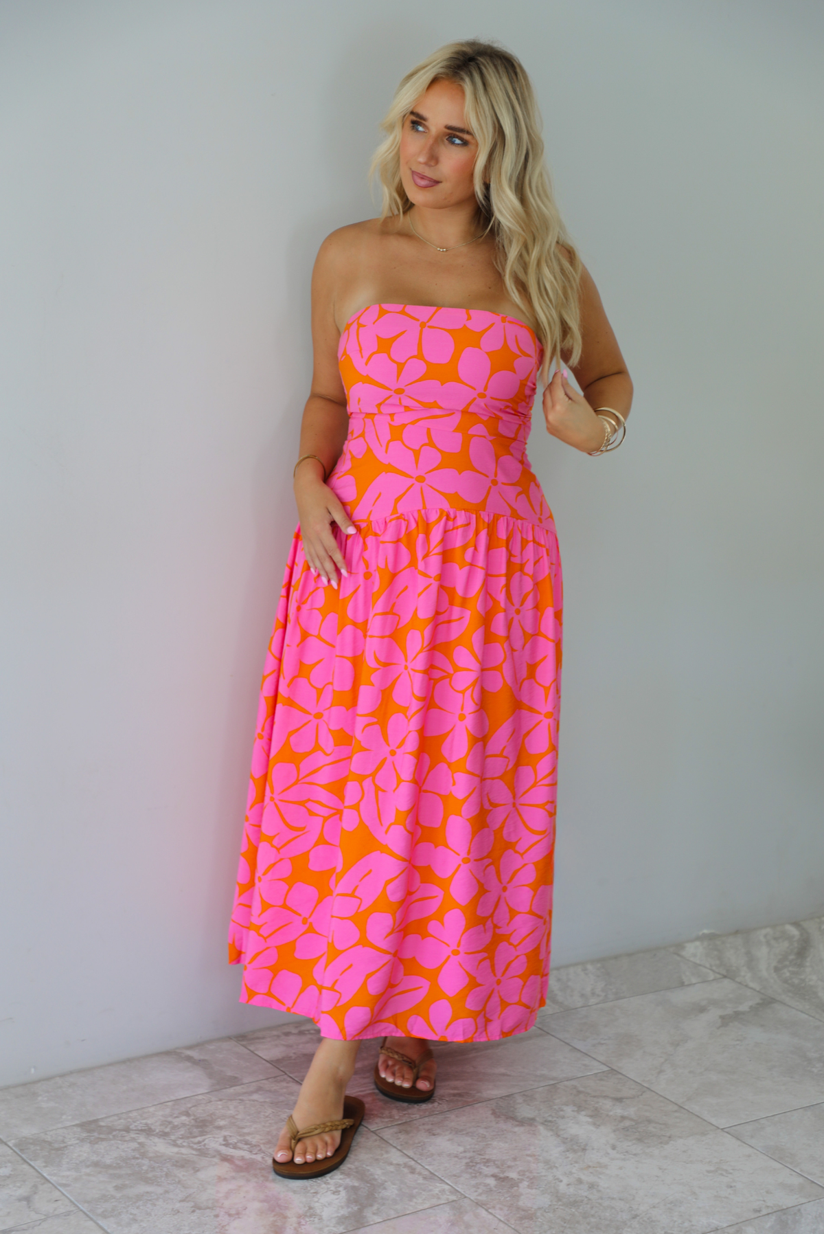 A woman modeling a strapless midi dress with a vibrant pink and orange floral pattern, standing indoors agains a white wall with white marble flooring. 