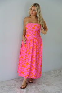 A woman modeling a strapless midi dress with a vibrant pink and orange floral pattern, standing indoors agains a white wall with white marble flooring. 