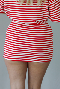Woman wearing a red and white striped set featuring an oversized top and mini skirt indoors against a white wall. The photo shows a closeup of the back of the skirt. 