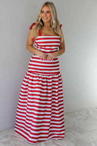 Woman wearing a red and white striped two piece set featuring a cropped top and maxi length, high waisted skirt. The photo is taken indoors against a white wall with white marble flooring. 