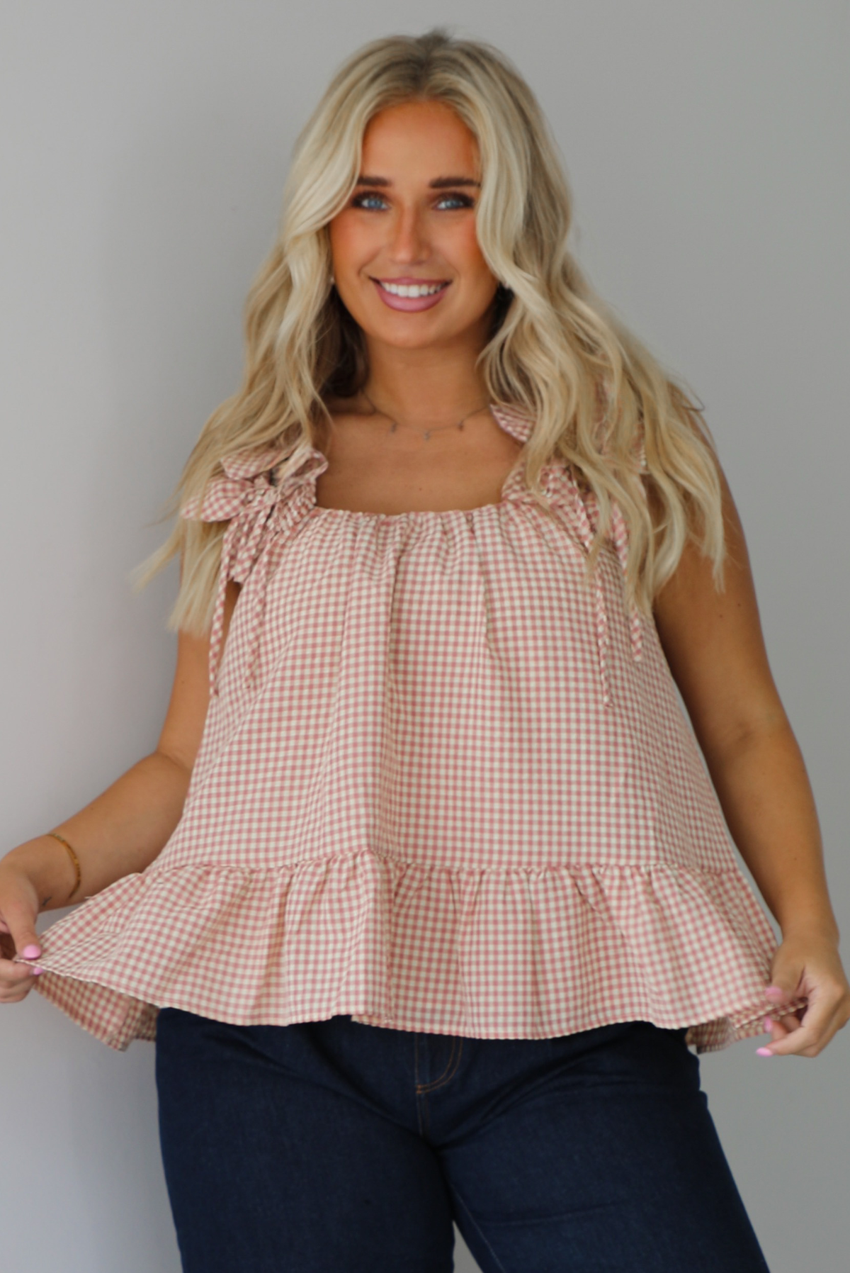 A woman wearing a light maroon and cream gingham flowy top with floral details at the straps of the tank, paired with dark denim jeans, standing indoors against a white wall.