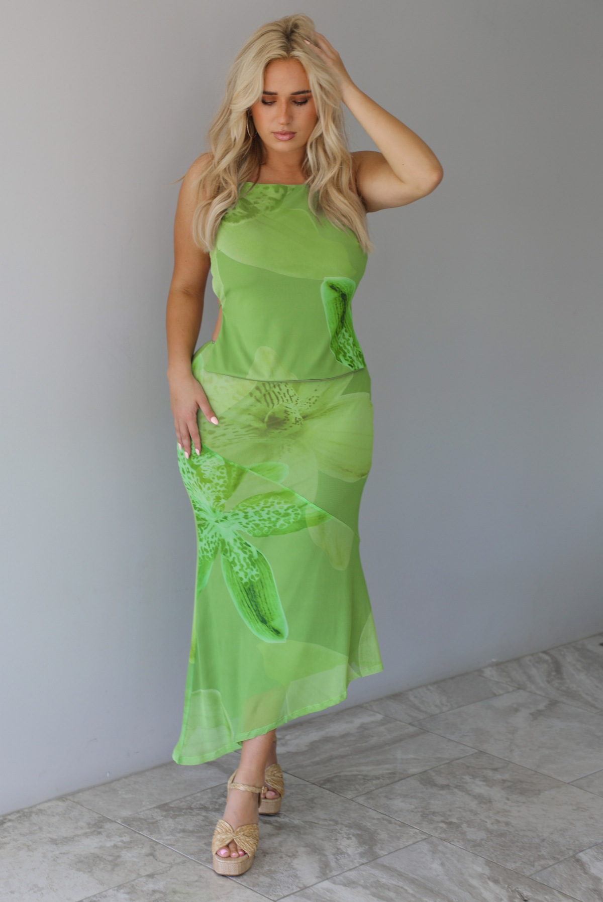 Woman wearing a green tropical print set featuring a backless tank and high waisted, asymmetrical hem midi skirt indoors against a white wall with white marble flooring. 