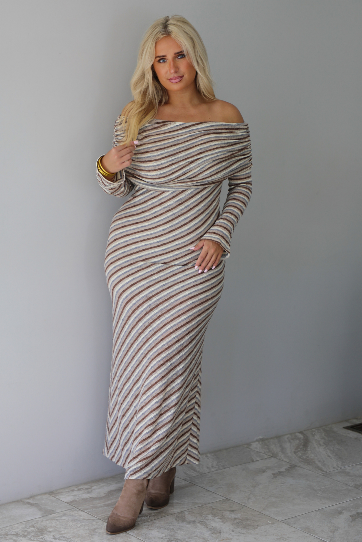 Woman wearing a grey, brown, muted blue, and off white striped fitted maxi dress that has a folded down design at the shoulders and bodice and long sleeves, standing indoors against a white wall with white marble flooring. 