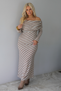 Woman wearing a grey, brown, muted blue, and off white striped fitted maxi dress that has a folded down design at the shoulders and bodice and long sleeves, standing indoors against a white wall with white marble flooring. 