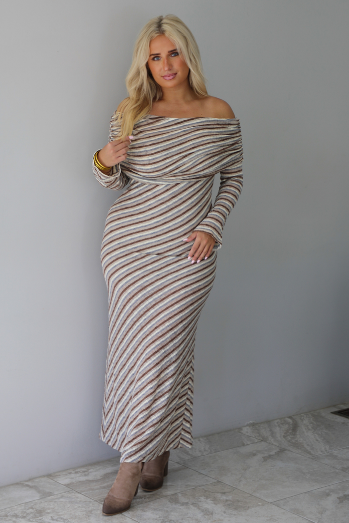 Woman wearing a grey, brown, muted blue, and off white striped fitted maxi dress that has a folded down design at the shoulders and bodice and long sleeves, standing indoors against a white wall with white marble flooring. 