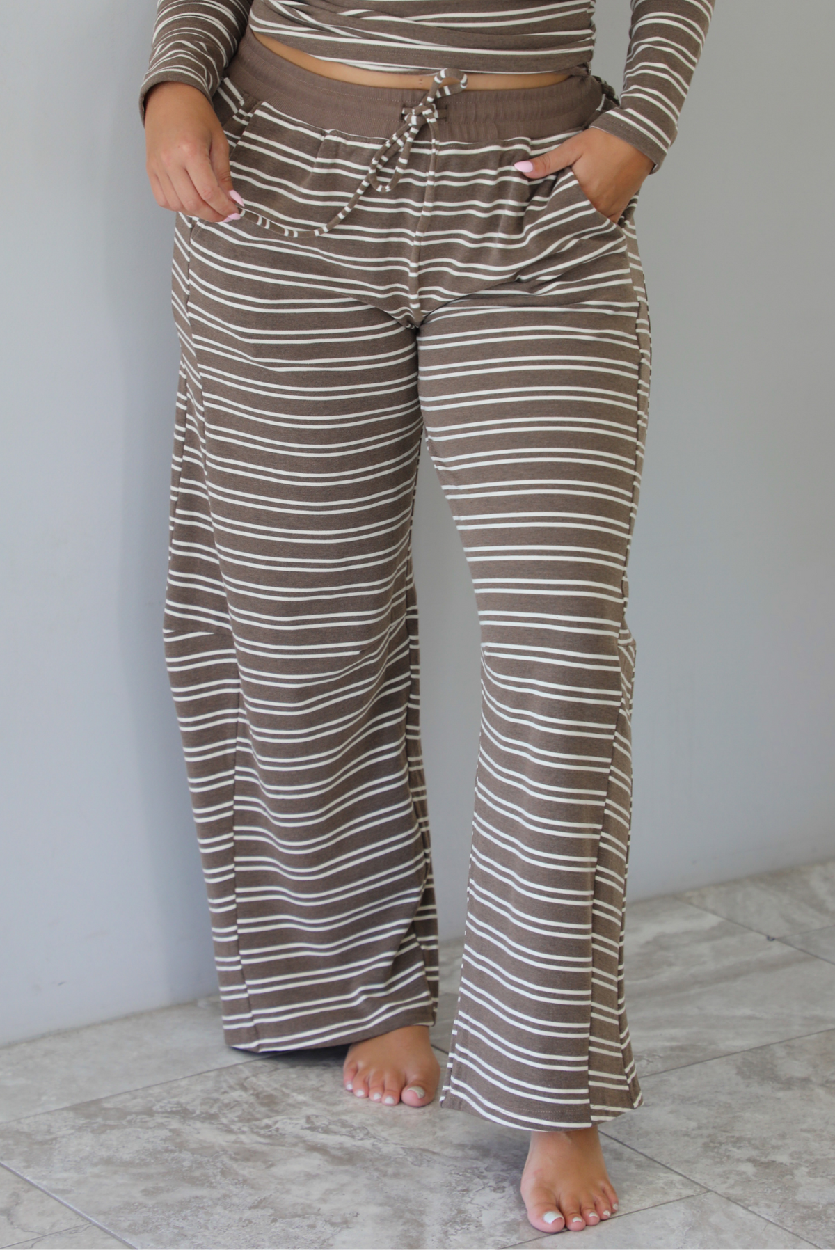 A woman wearing a brown with cream striped matching two piece set consisting of a long sleeved, off the shoulder top and high waisted pants, standing indoors against a white wall with white marble flooring. The photo shows a closeup of the pants, showing the elastic and drawstring waistline and functional pocket on either side. 