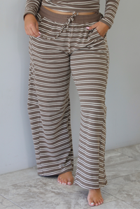 A woman wearing a brown with cream striped matching two piece set consisting of a long sleeved, off the shoulder top and high waisted pants, standing indoors against a white wall with white marble flooring. The photo shows a closeup of the pants, showing the elastic and drawstring waistline and functional pocket on either side. 