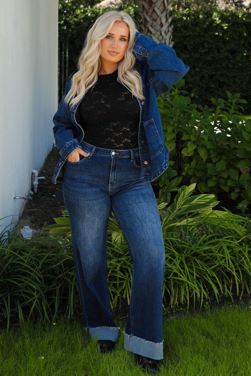 Woman model wearing a dark denim button up jacket with a black lace top,  and dark denim jeans outdoors. 