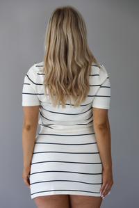 Woman wearing a white and black striped set with functional button closures on the top. The photo is taken indoors against a white wall and shows the back of the set. 