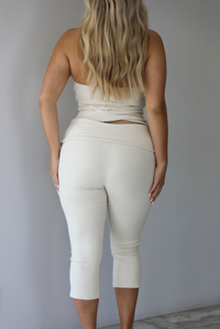 Woman wearing a cream two piece set featuring a strapless tube top and capri pants that fold over at the waist indoors against a white wall. The photo shows a closeup of the back of the set. 