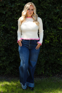 Woman model wearing a cream knit top with pink trim and dark denim jeans. The photo is taken outdoors against greenery. 