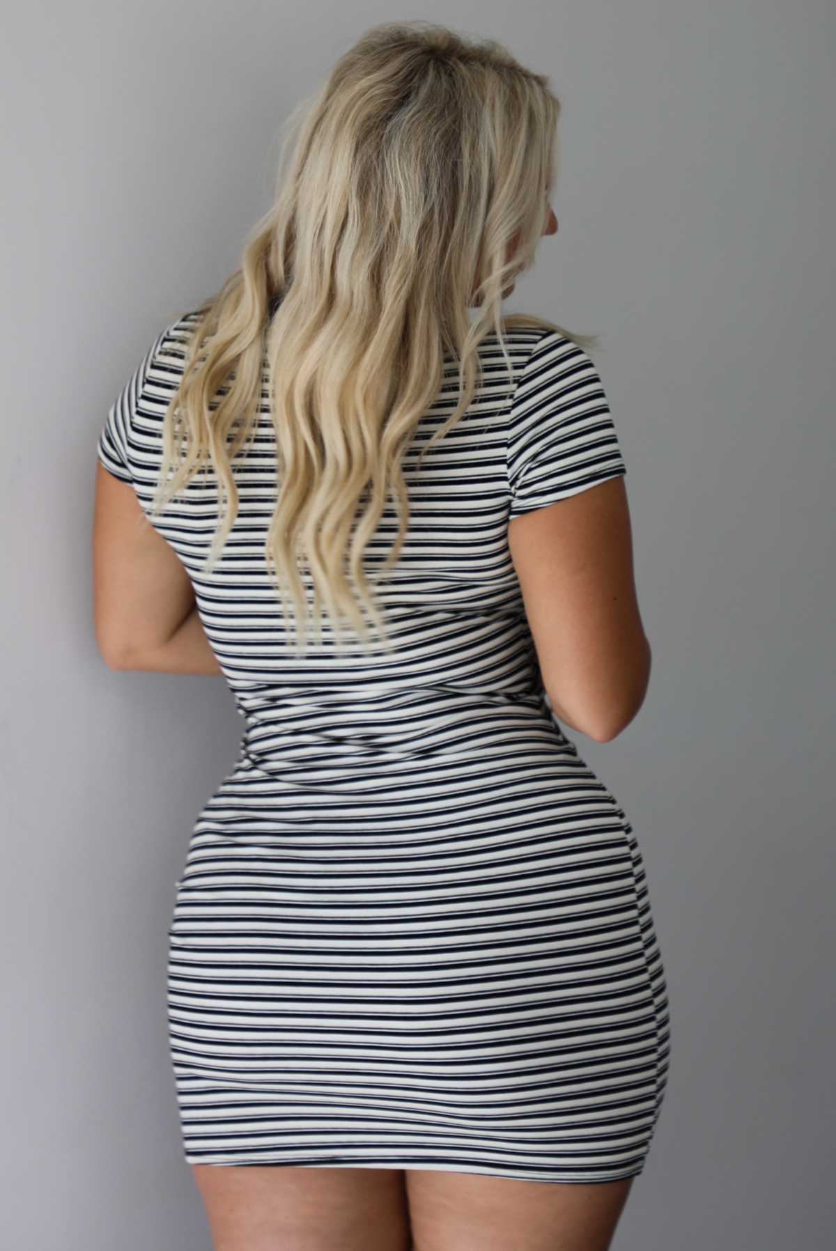 A woman shown from behind wearing a black and white striped fitted mini t-shirt dress featuring a high neckline, short sleeves, with a mini length, standing indoors against a white wall. 