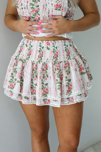 Woman wearing a white and pink floral two piece set featuring a strapless corset style top and an a ruffled skirt. The photo shows a closeup of the skirt, with its elastic waistline. The photo is taken indoors against a white wall.