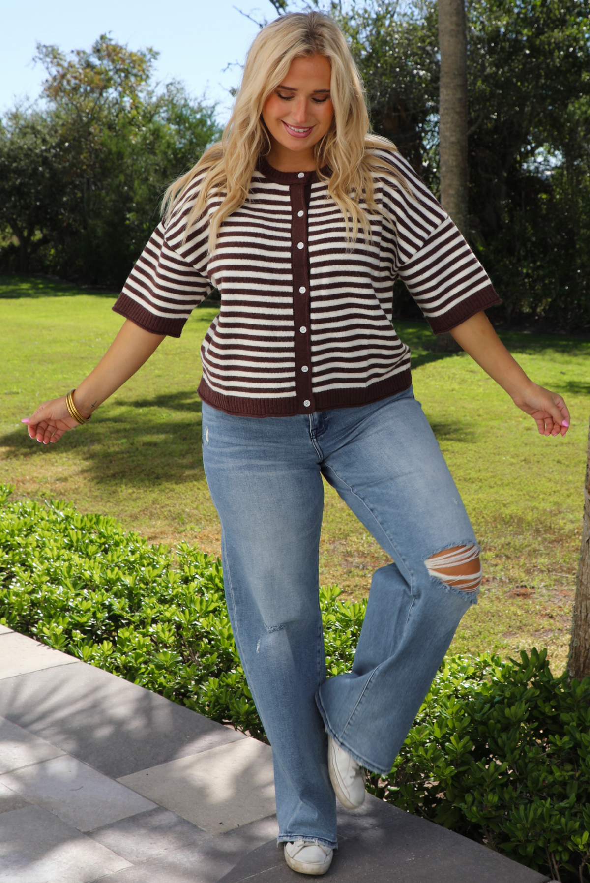 A woman wearing a brown and white striped knit button top cardigan top, paired with light wash denim distressed denim, standing outdoors on a stone path with palm trees and greenery in the background. 