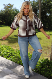 A woman wearing a brown and white striped knit button top cardigan top, paired with light wash denim distressed denim, standing outdoors on a stone path with palm trees and greenery in the background. 