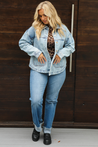 Woman model wearing an oversized, acid washed, light denim jacket, paired with a sheer mesh leopard print top, medium wash denim jeans, and black shoes. The photo is taken outdoors against a dark brown door. 