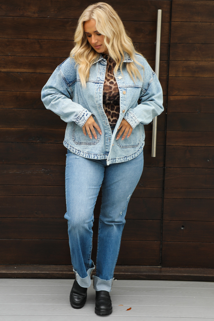 Woman model wearing an oversized, acid washed, light denim jacket, paired with a sheer mesh leopard print top, medium wash denim jeans, and black shoes. The photo is taken outdoors against a dark brown door. 