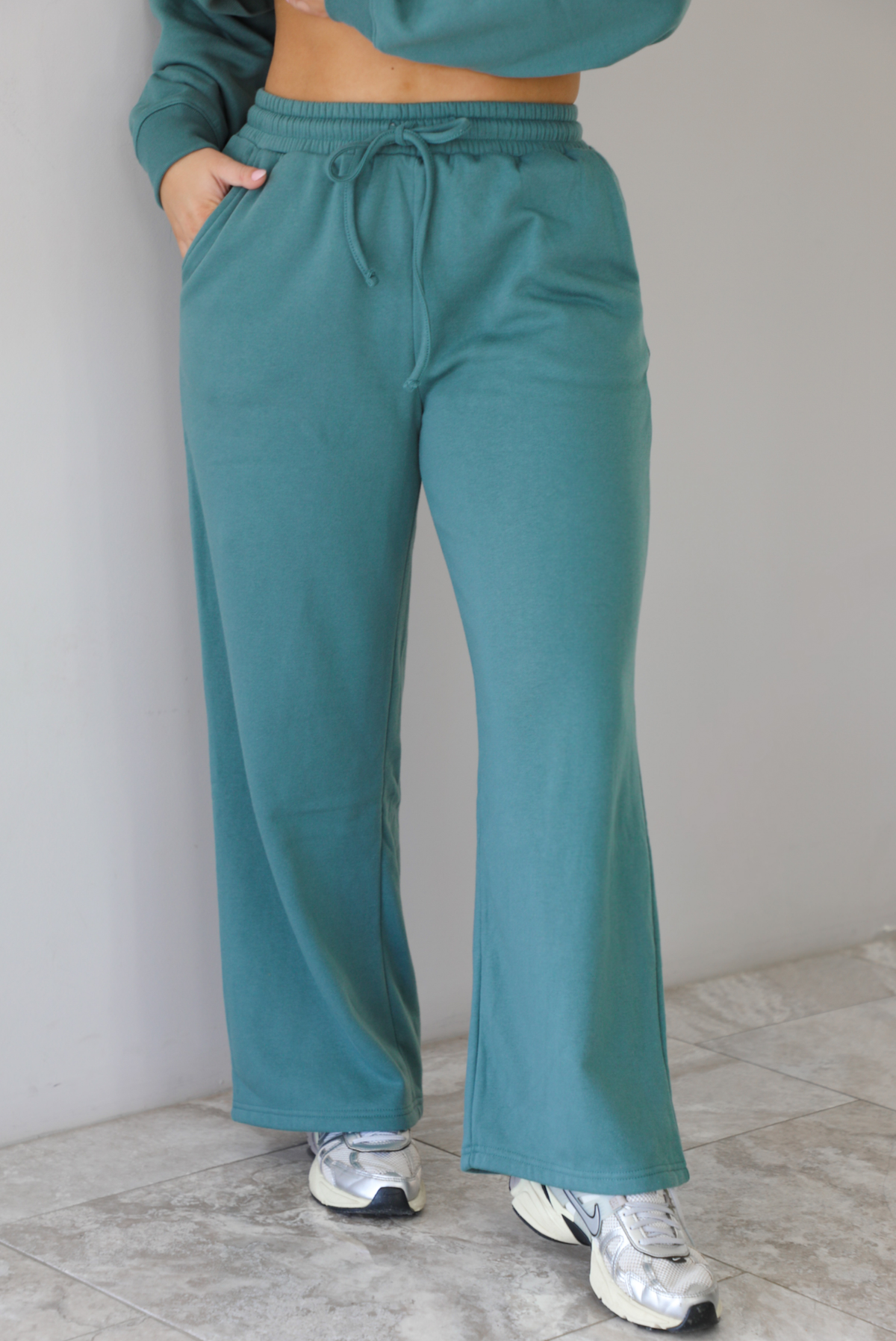Woman wearing a sage two piece sweat-set. The set features a slightly oversized sweater and high waisted sweat pants that have an elastic and drawstring waistline with pockets.The photo shows a closeup of the pants. The photo is taken indoors against a white wall with white marble flooring. 