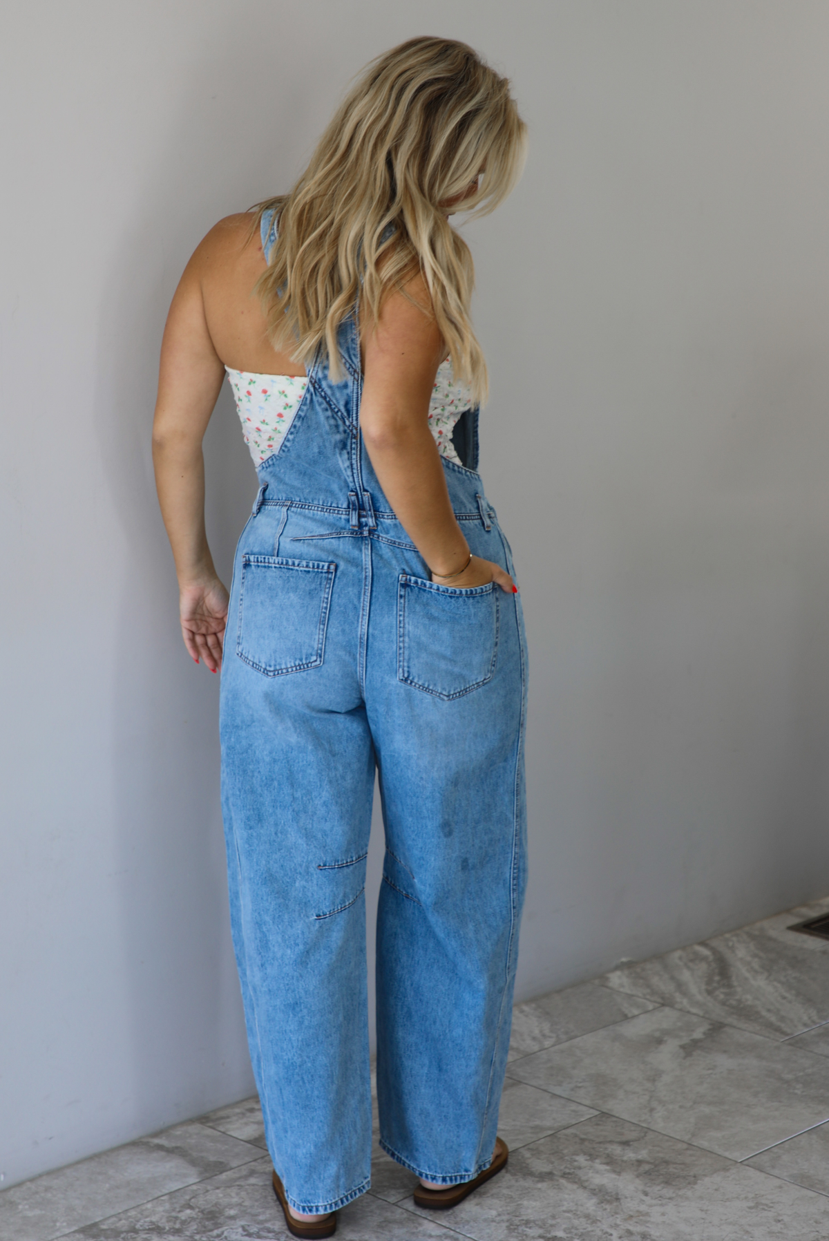 Woman wearing a pair of denim overalls with a white floral strapless top underneath indoors against a white wall with white marble flooring. The photo shows the back of the overalls. 