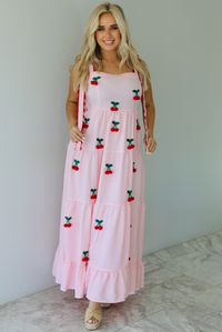 Woman wearing a pink and white gingham  maxi dress with thick straps, a fitted bodice, flowy skirt, and crocheted cherry details on the front indoors against a white wall with white marble flooring. 