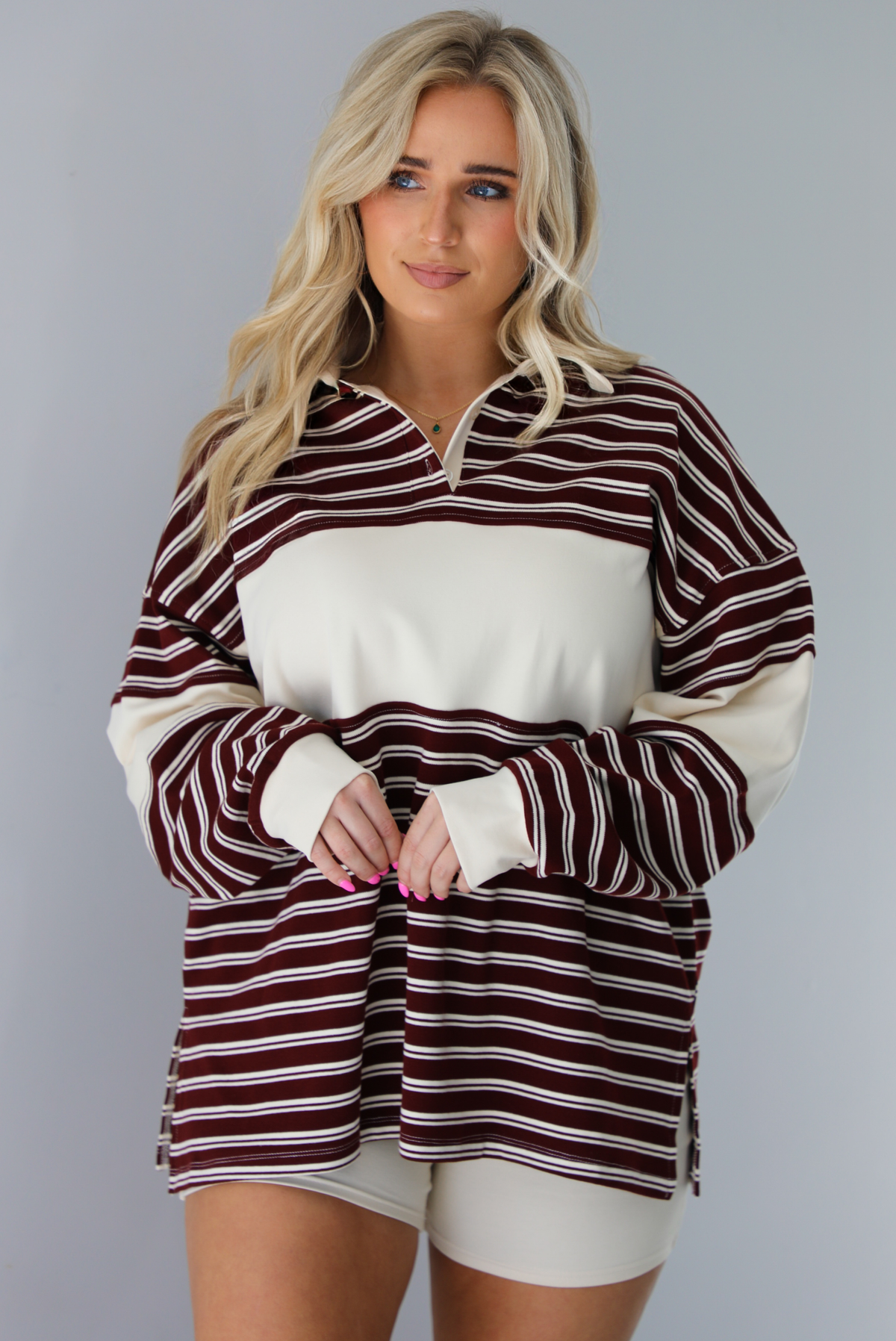 Woman wearing a burgundy and cream striped oversized collared top with cream colored biker shorts. The photo is taken indoors against a white wall.