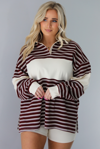 Woman wearing a burgundy and cream striped oversized collared top with cream colored biker shorts. The photo is taken indoors against a white wall.